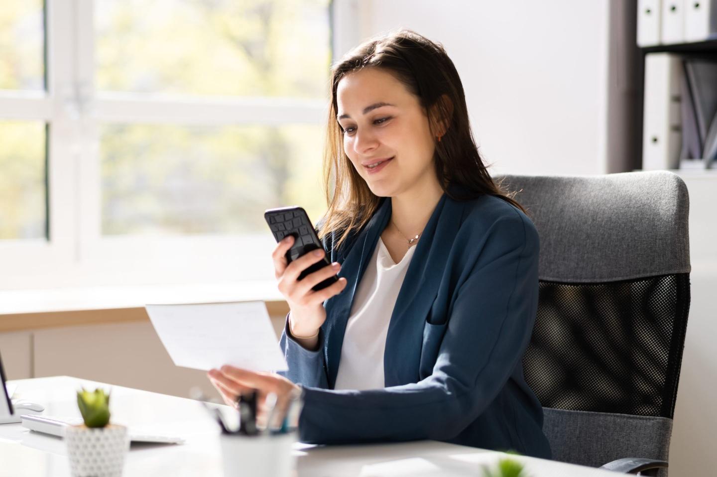 A woman dressed in business attire deposits a check using a cell phone.