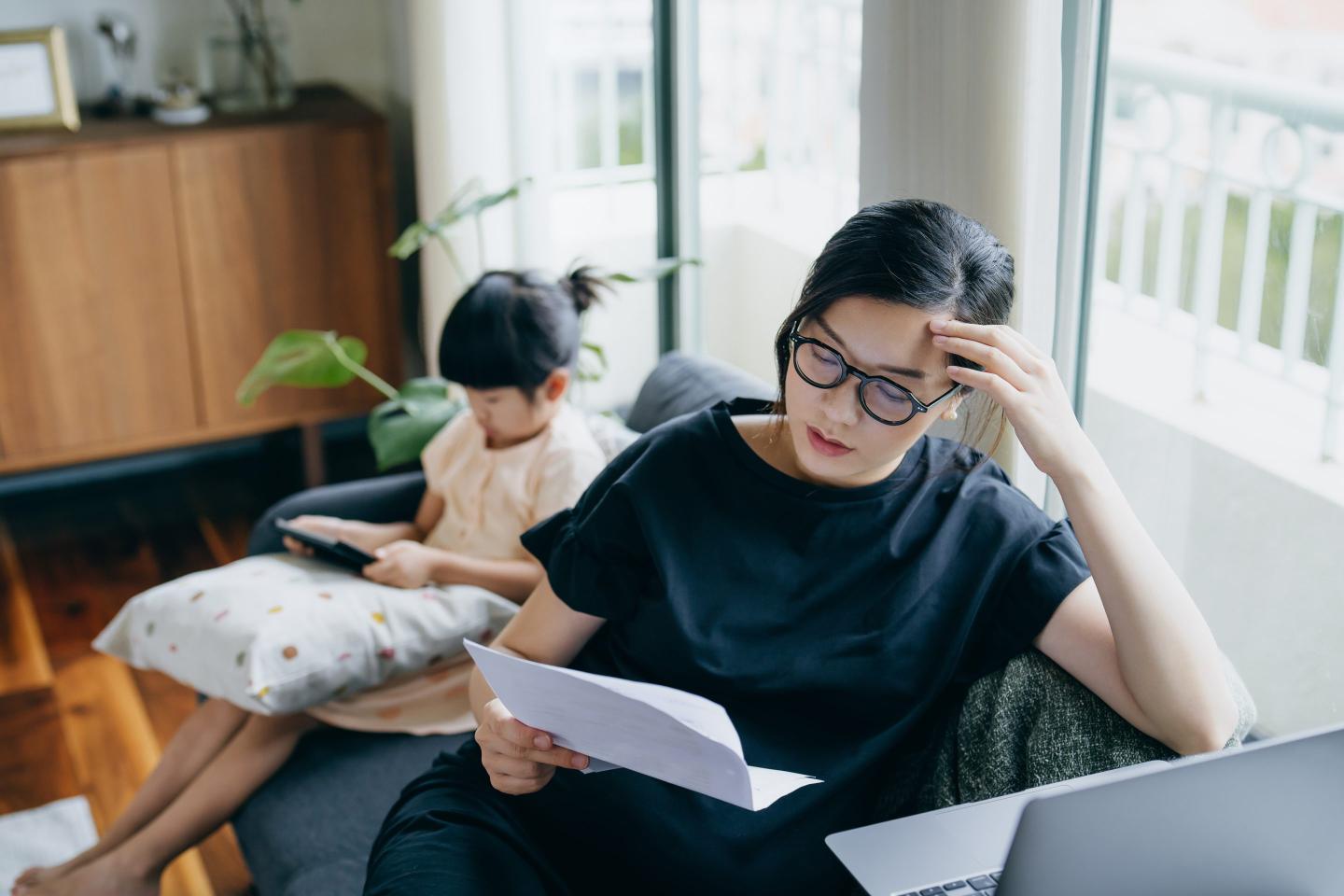 Stock photo of a young Asian woman looking stressed while managing her financial bills and tax documents, while her daughter uses a digital tablet in the background.