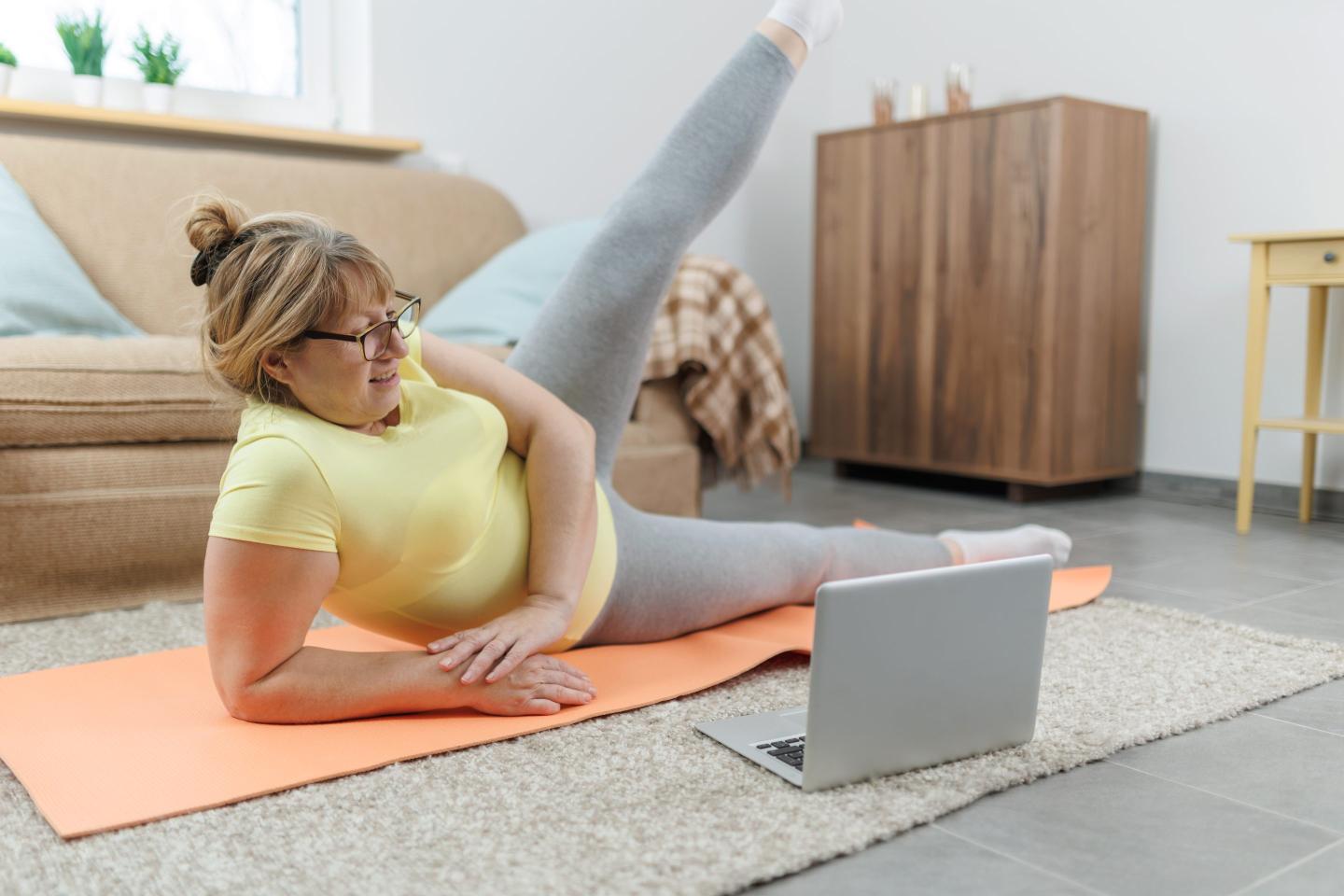 Mature woman exercising on the floor in front of laptop in the living room at home
