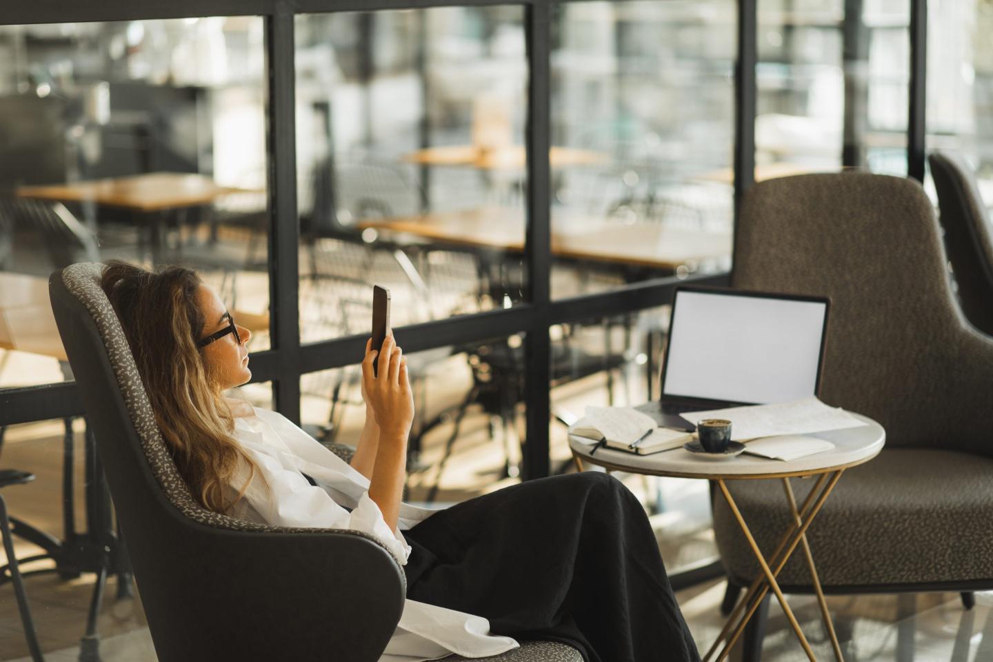 A woman reclines in an arm chair while looking at her phone. In front of her is a coffee table with a laptop, espresso drink, and notebook set up.