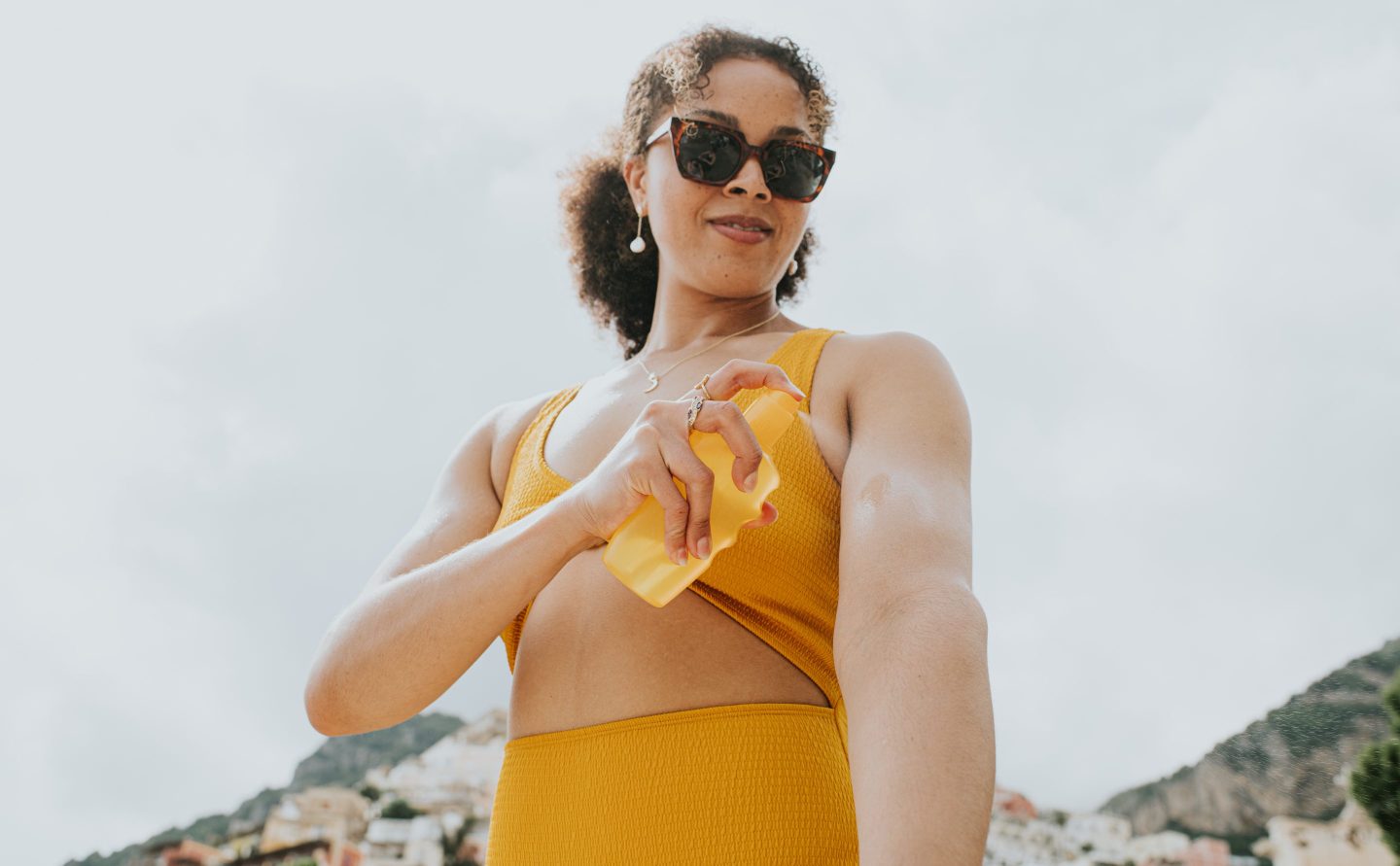 Waist up image of a woman wearing sunglasses, spraying suncream on to her arm.