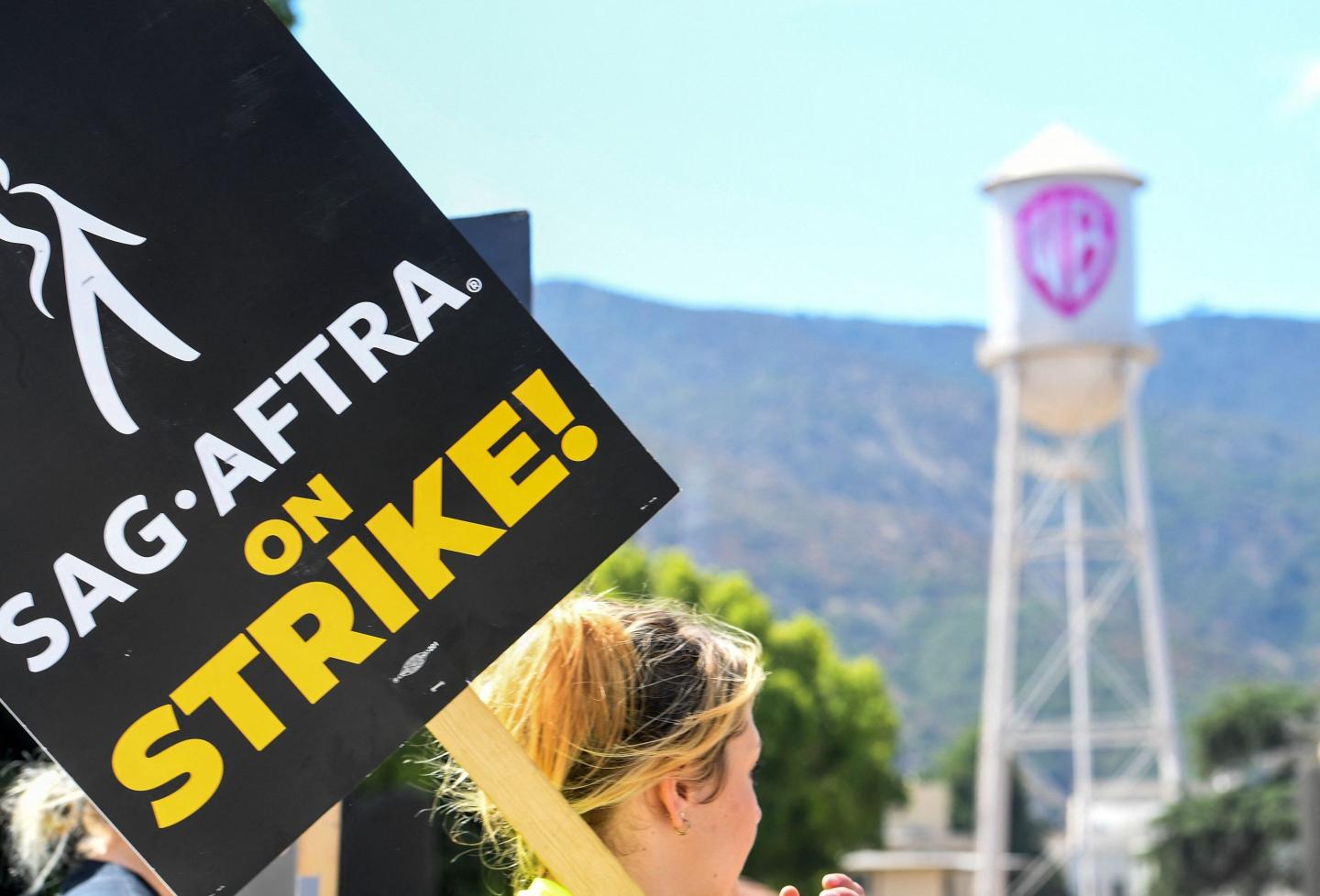 Picketer carrying a sign reading "SAG-AFTRA on strike!" with the Warner Bros. water tower in the background.