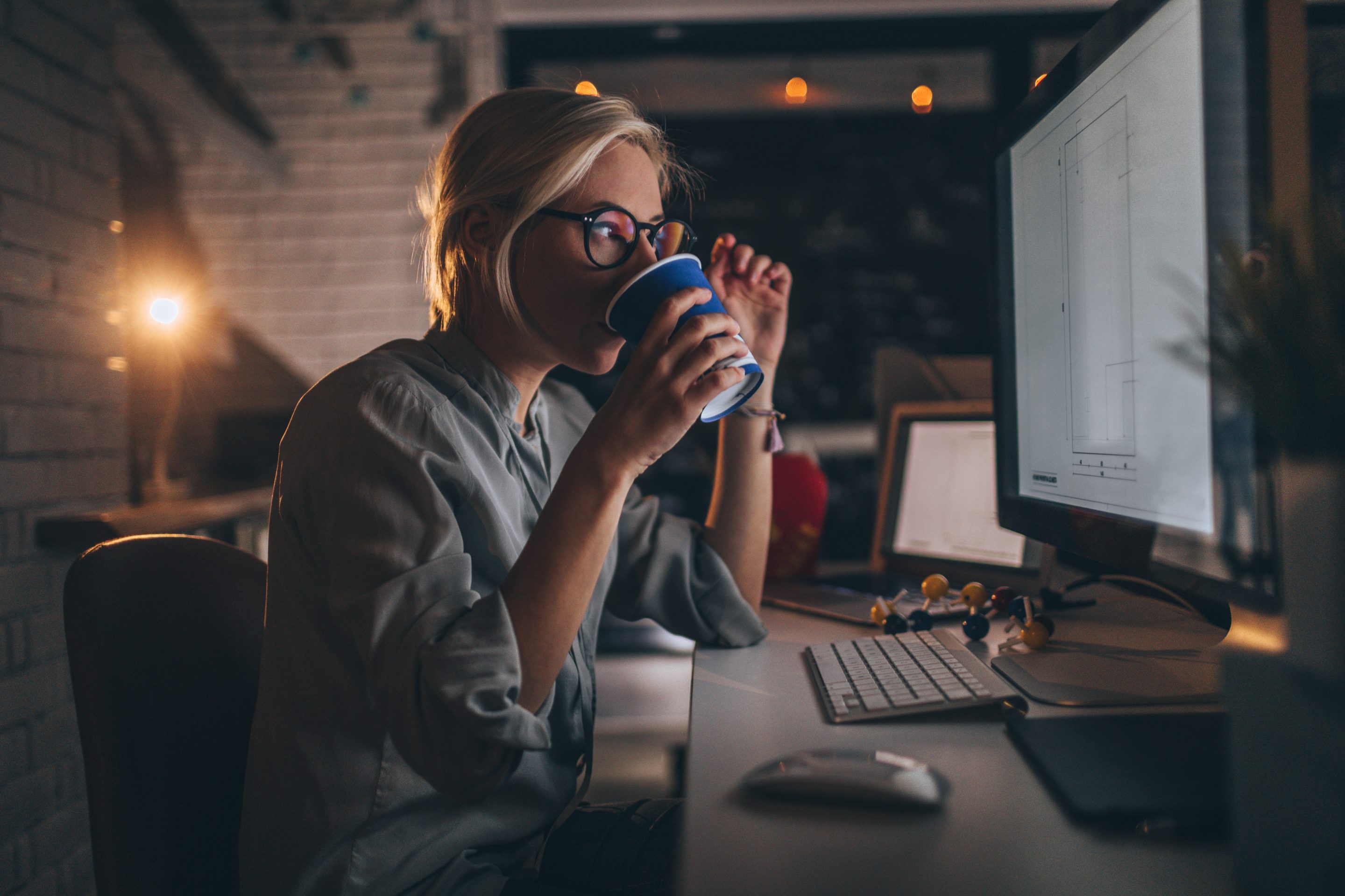 Young woman sitting at her computer, having some coffee to help her through a late night shift