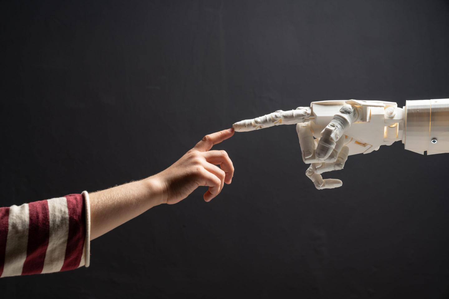 A child’s finger and a humanoid robotic finger touch in front of a black background resembling Michaelangelo's painting, "The Creation of Adam"