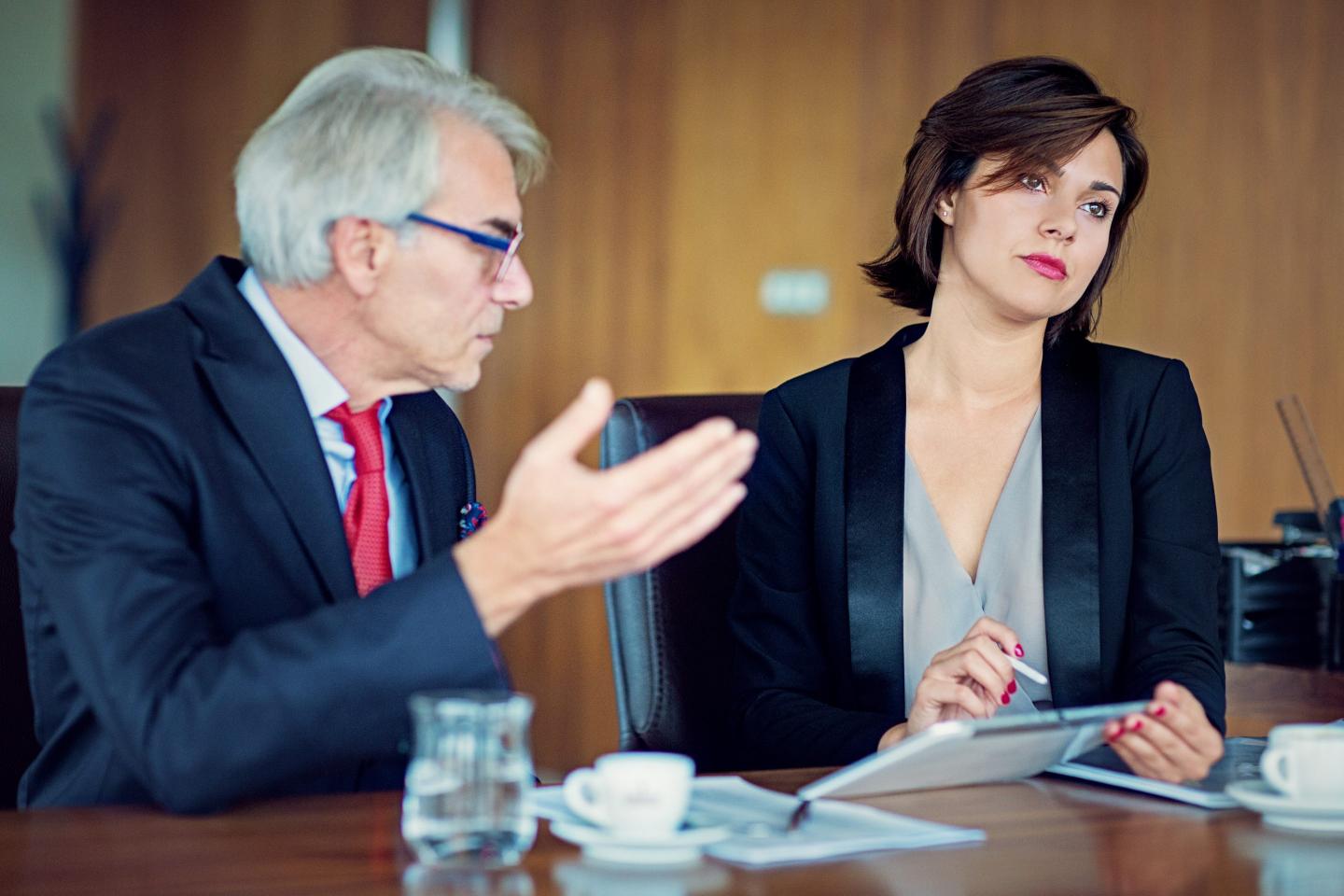 An older businessman speaks to a younger businesswoman who is rolling her eyes.