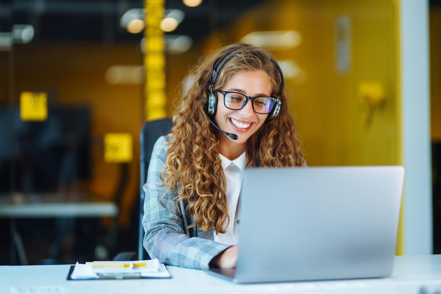 A young business woman sits in an office, using a laptop and wearing a headset.