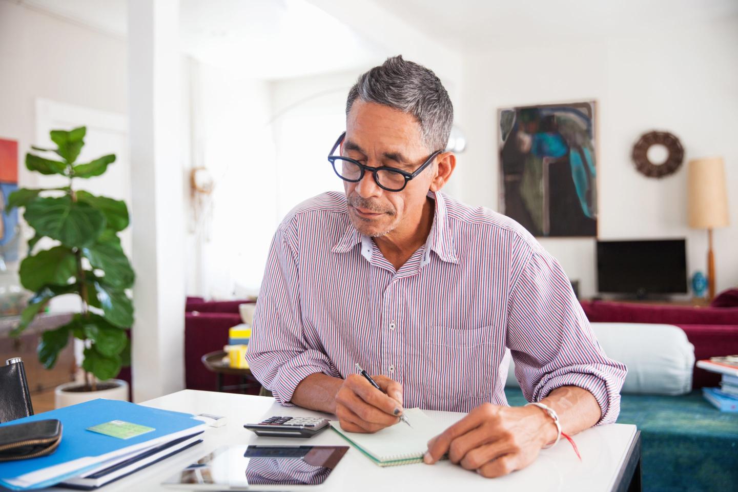 Stock image of a man sitting at a desk looking at a calculator and writing notes on a notepad.