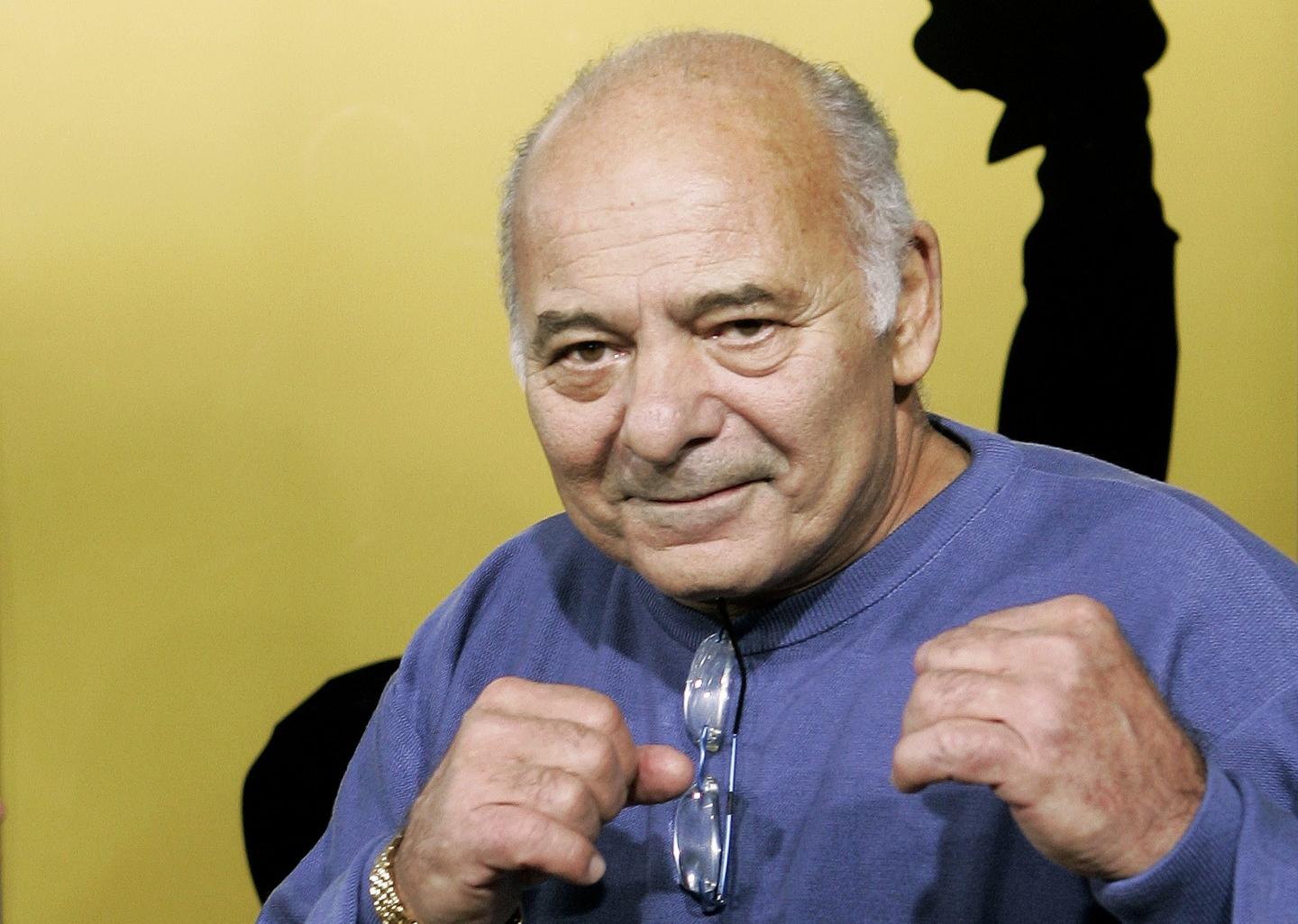 Burt Young wearing a blue shirt and posed with his hands raised in fists as though he's boxing