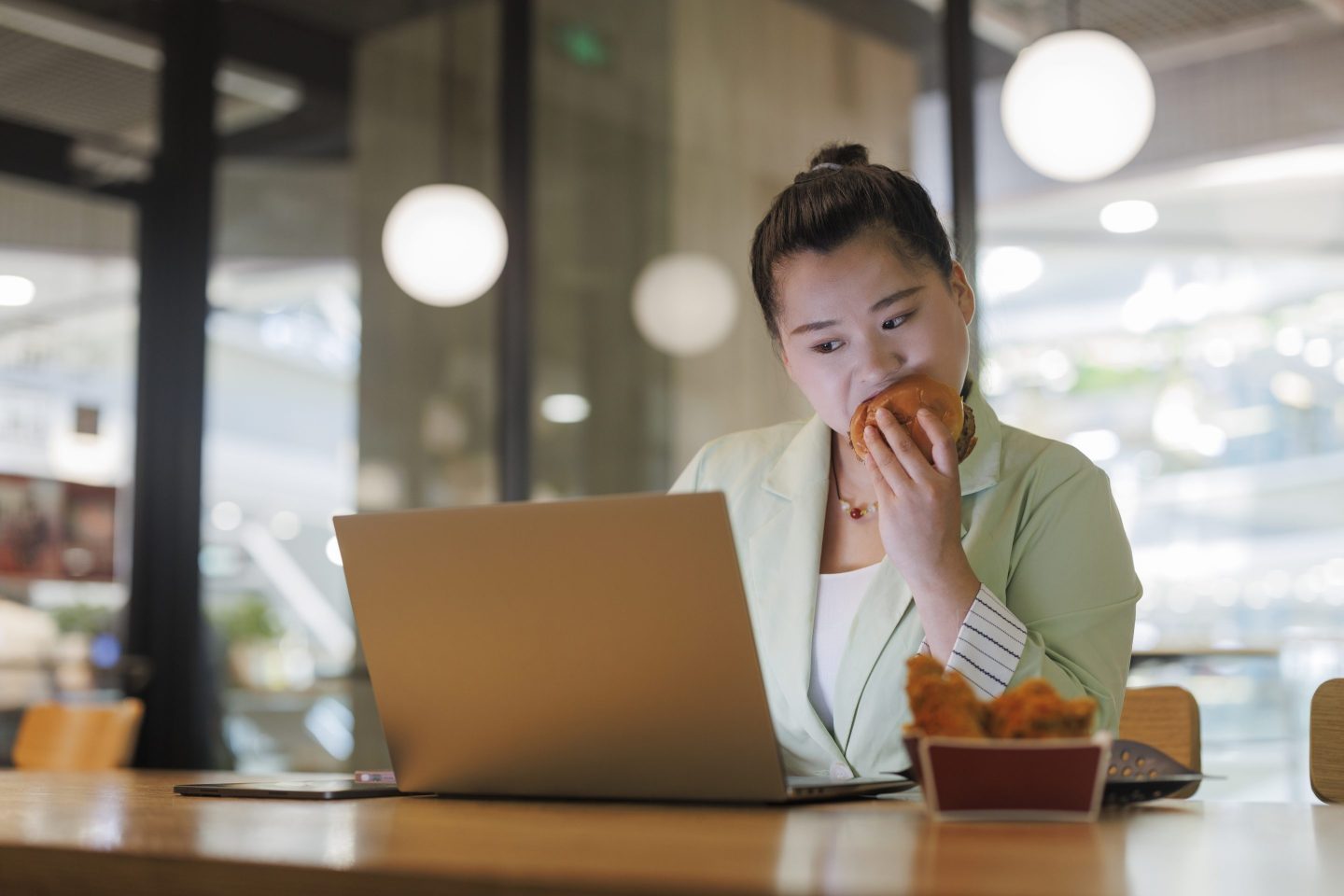 A woman eats a burger while working on her laptop.
