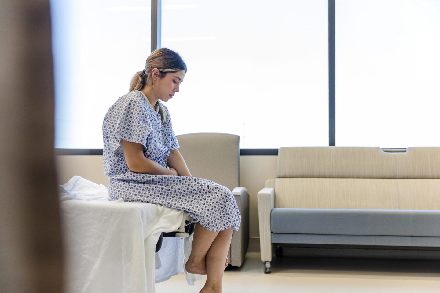 Anxious, sad, young woman wearing hospital gown looks down.