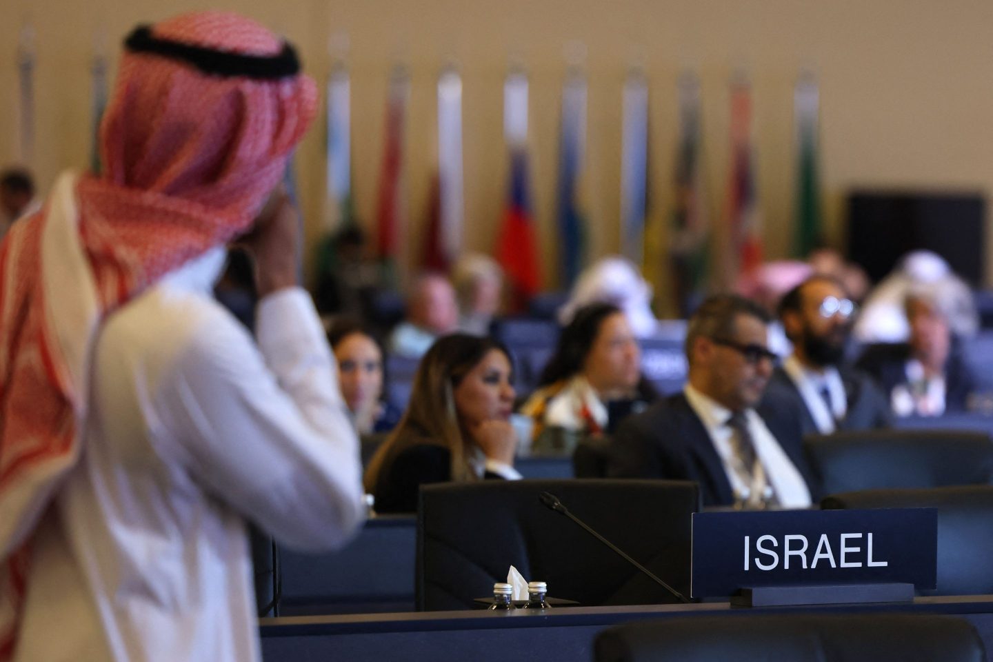 A Saudi Arabian man dressed in a traditional thobe stands in front of a sign the reads Israel in a large conference hall.