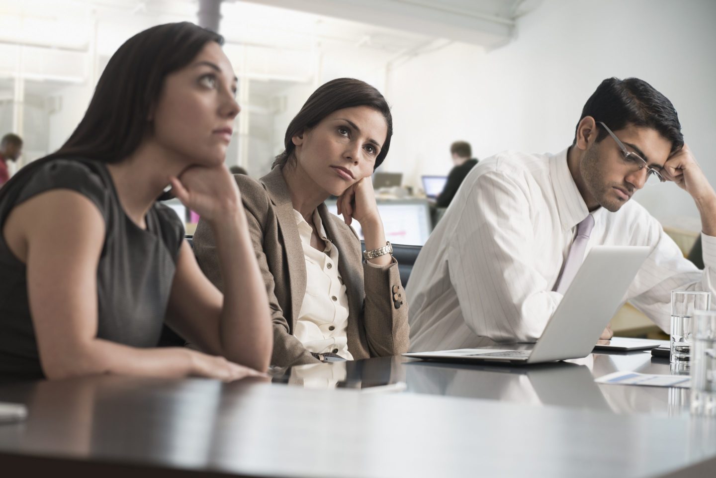 Three office workers, two women and a man, look bored and annoyed in a meeting.