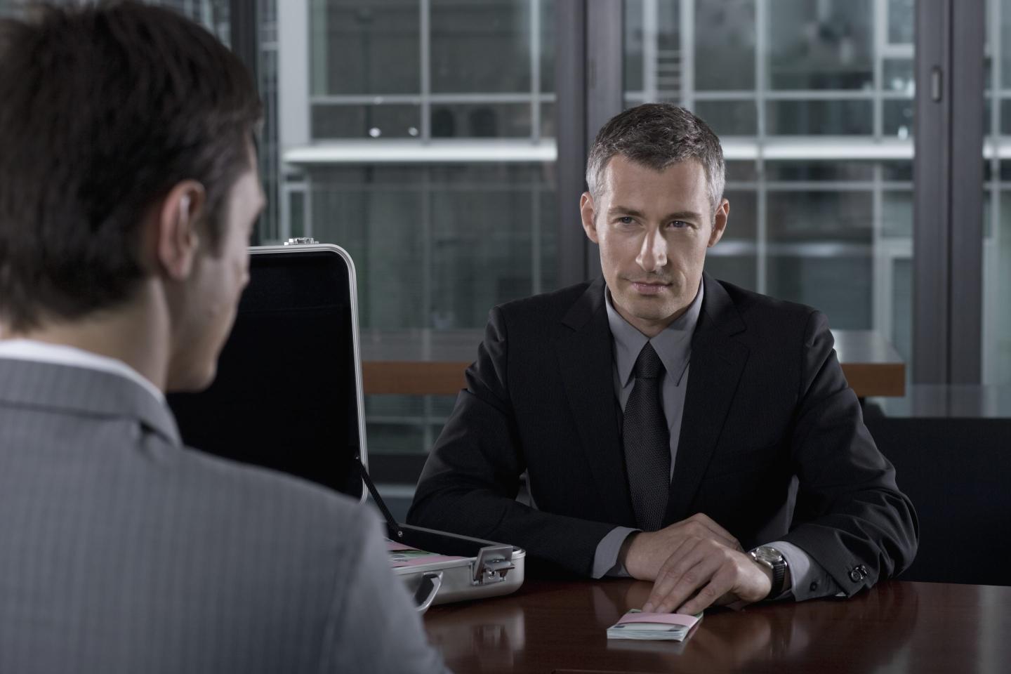 A business man bribing another business man by sliding a stack of money across a desk.