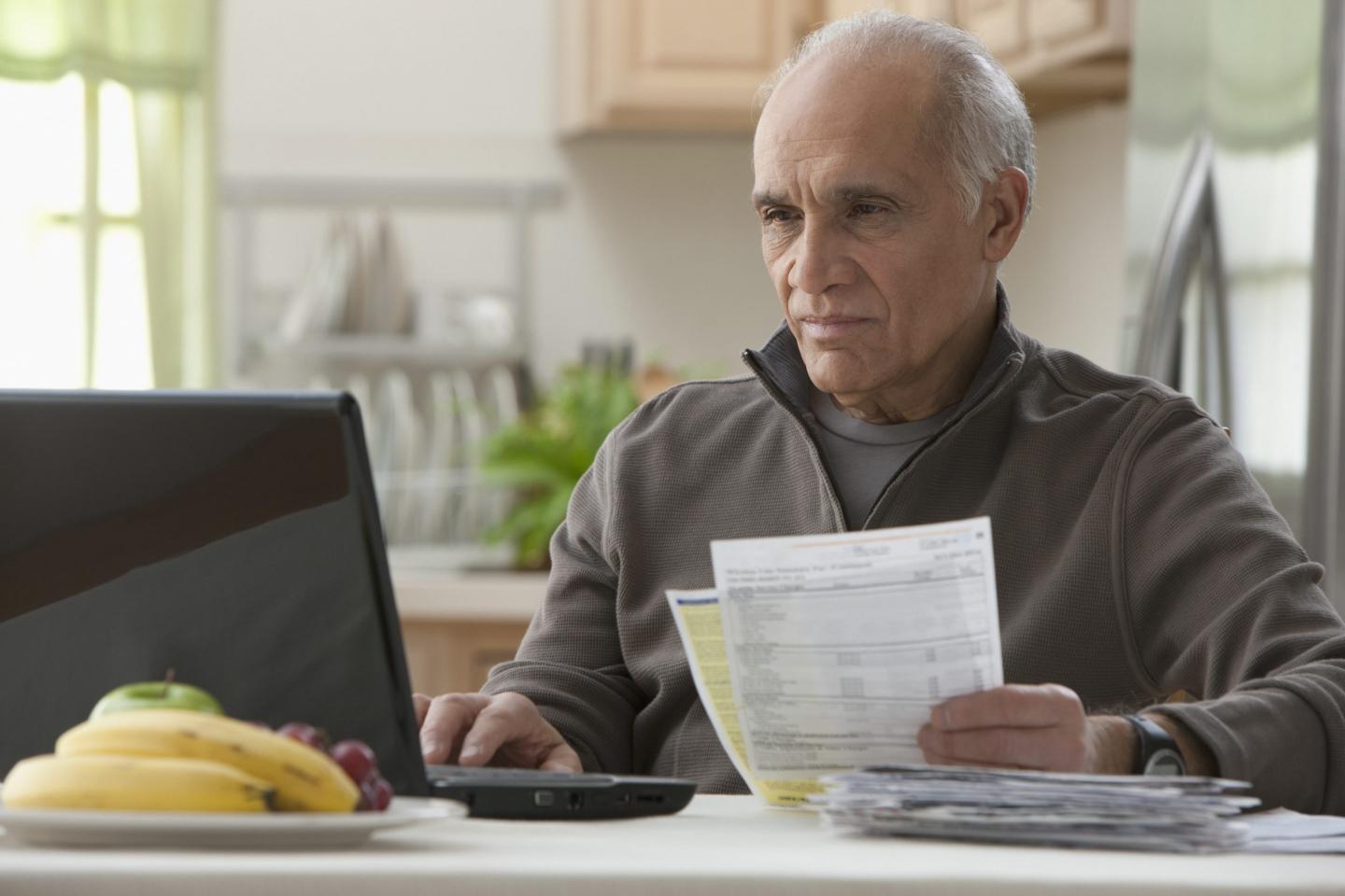 A middle-aged man sits in front of a laptop, holding bills