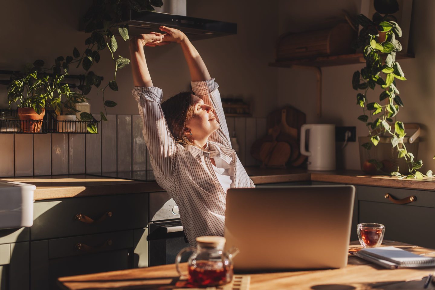 Happy relaxed young woman sitting in her kitchen with a laptop in front of her stretching her arms above her head and looking out of the window with a smile.