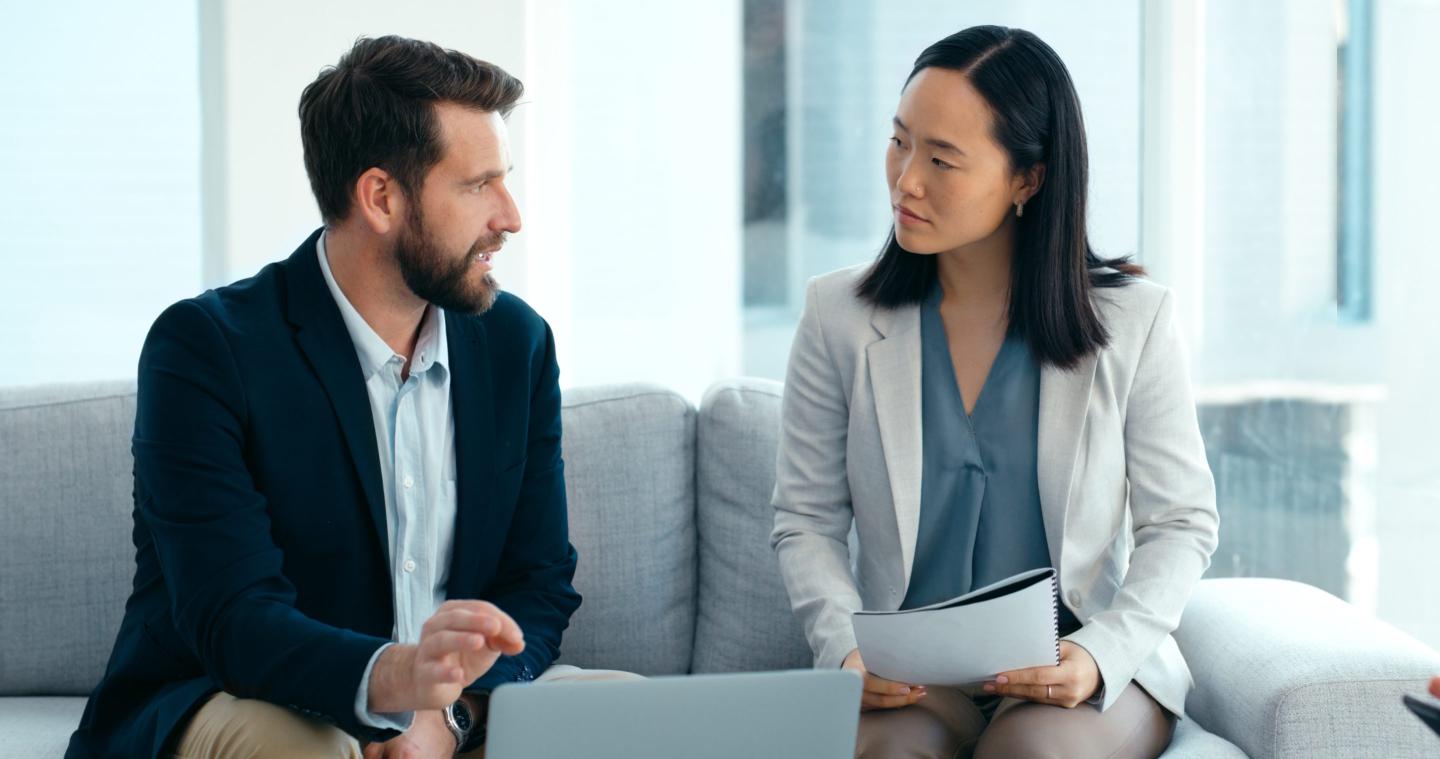A man and woman, both wearing business attire, sit on a couch in an office while having a work-related discussion.