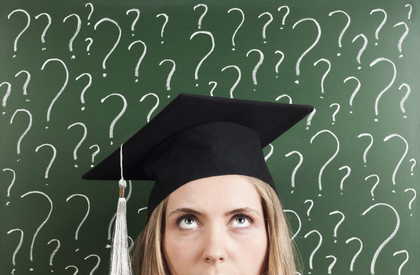 A young woman student wearing a graduation cap in front of a chalkboard with question marks written on it.