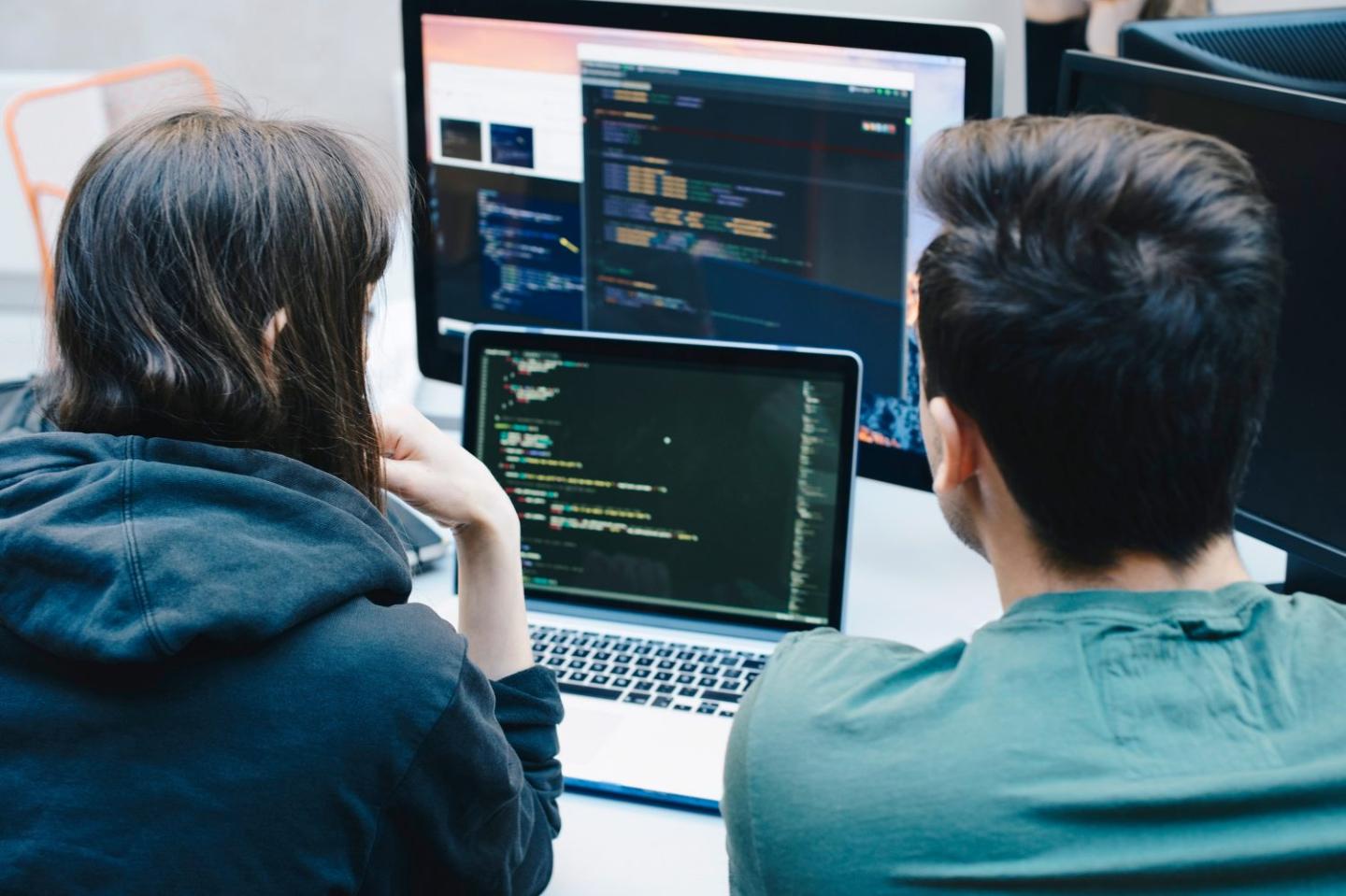 Photo of the rear view of two students looking at a laptop and computer monitor studying cybersecurity.