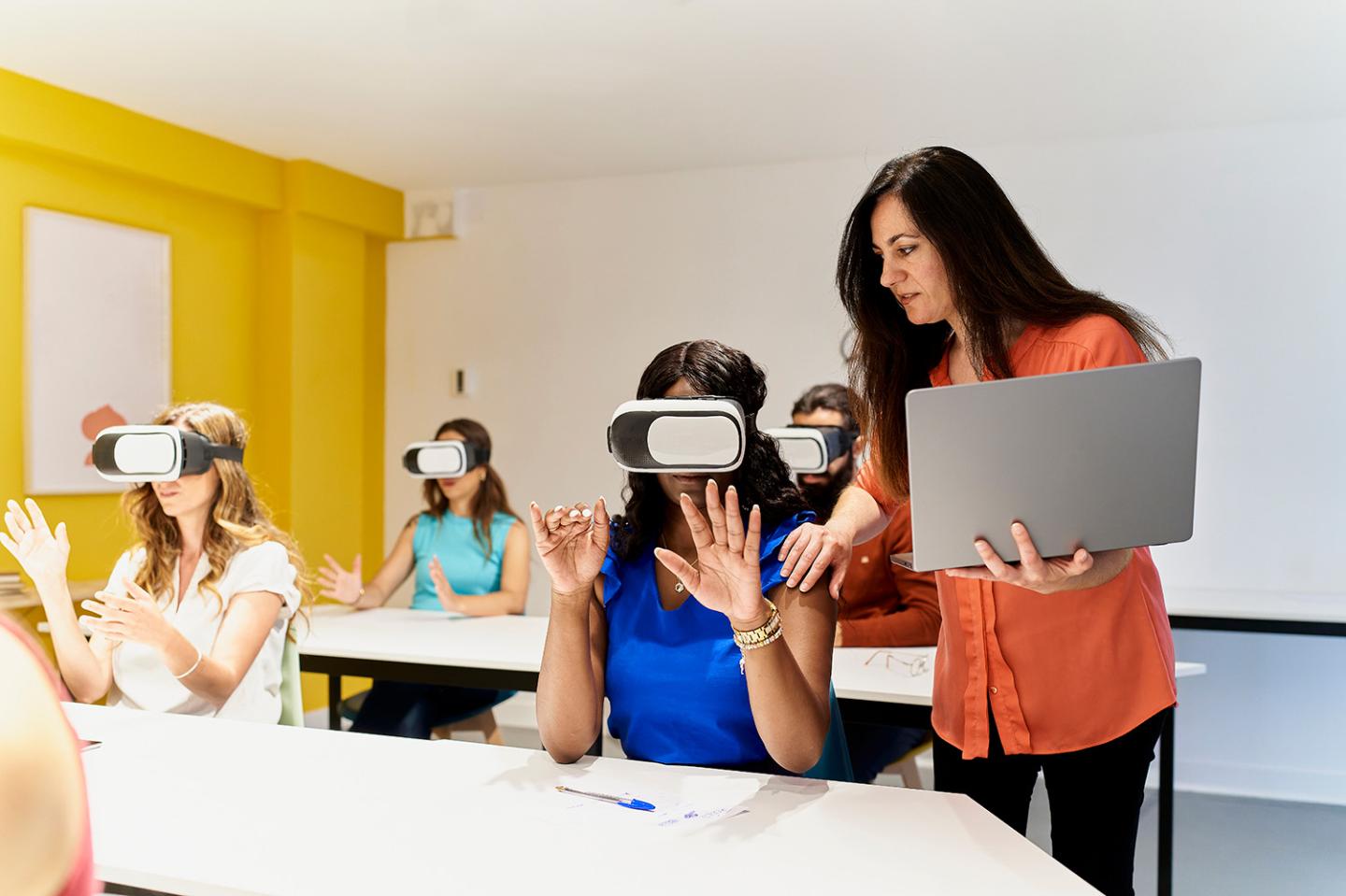 teacher in classroom holding laptop standing next to black female student with hand on her shoulder while instructing her on how to use virtual reality goggles