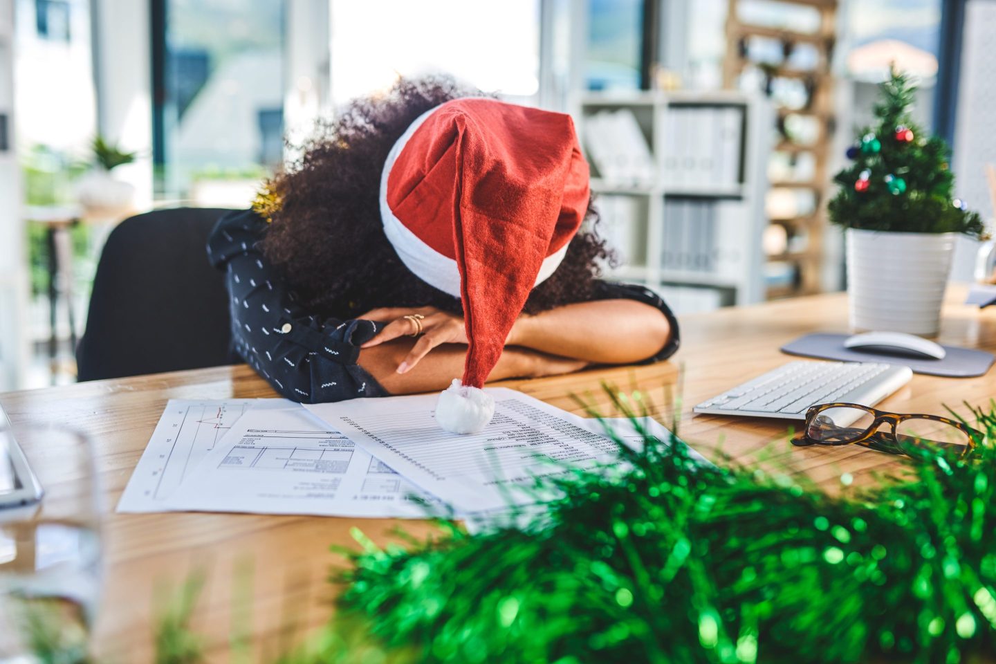 A young businesswoman wearing a Santa hat slumps over her desk, covered in paperwork.