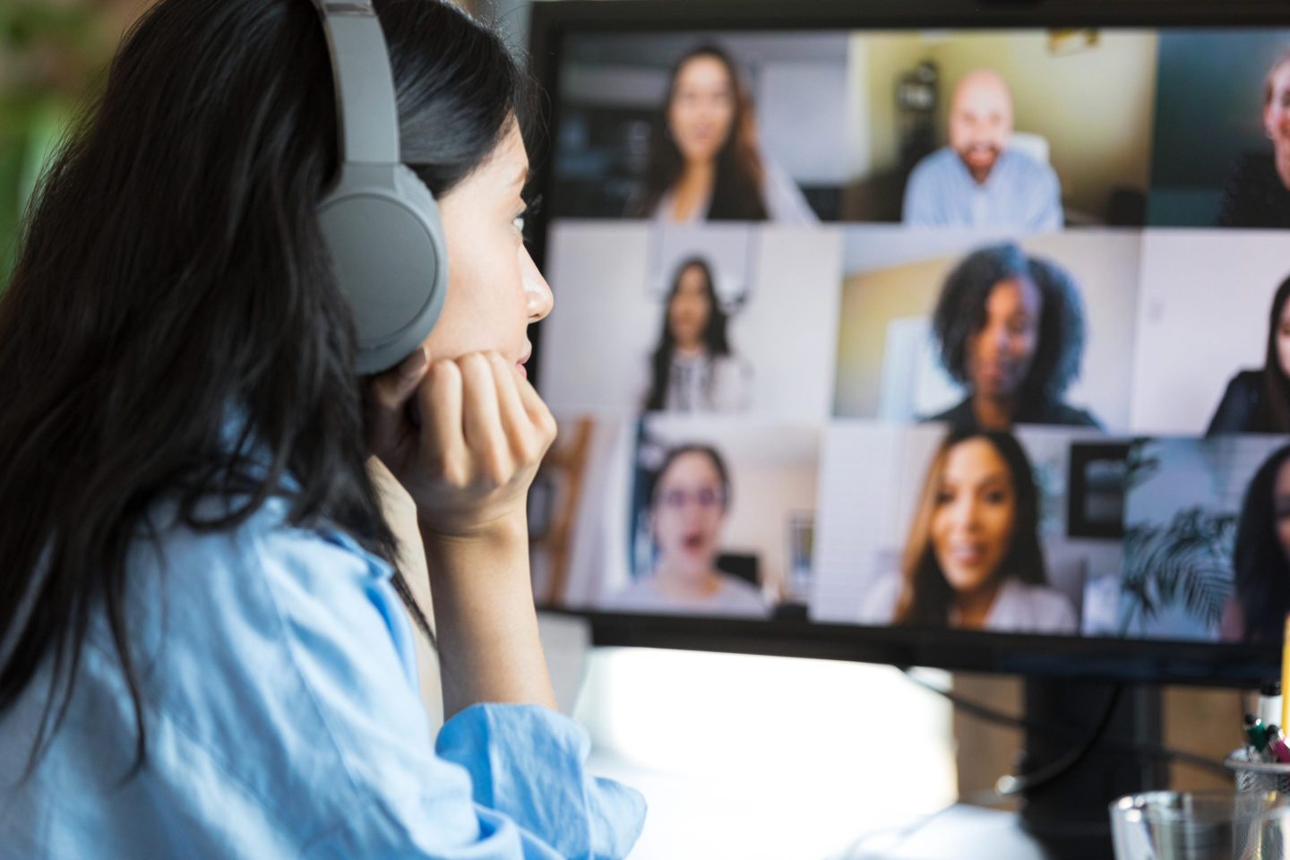A young woman wears Bluetooth headphones and rests her chin on her hand as she sits in front of a computer screen displaying a virtual meeting. She looks bored.