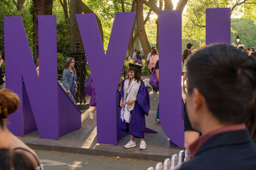 A New York University (NYU) graduate wearing a cap and gown takes photos near a large NYU sign in Washington Square Park, as seen in May 2022 in New York City. (Photo by Alexi Rosenfeld/Getty Images)