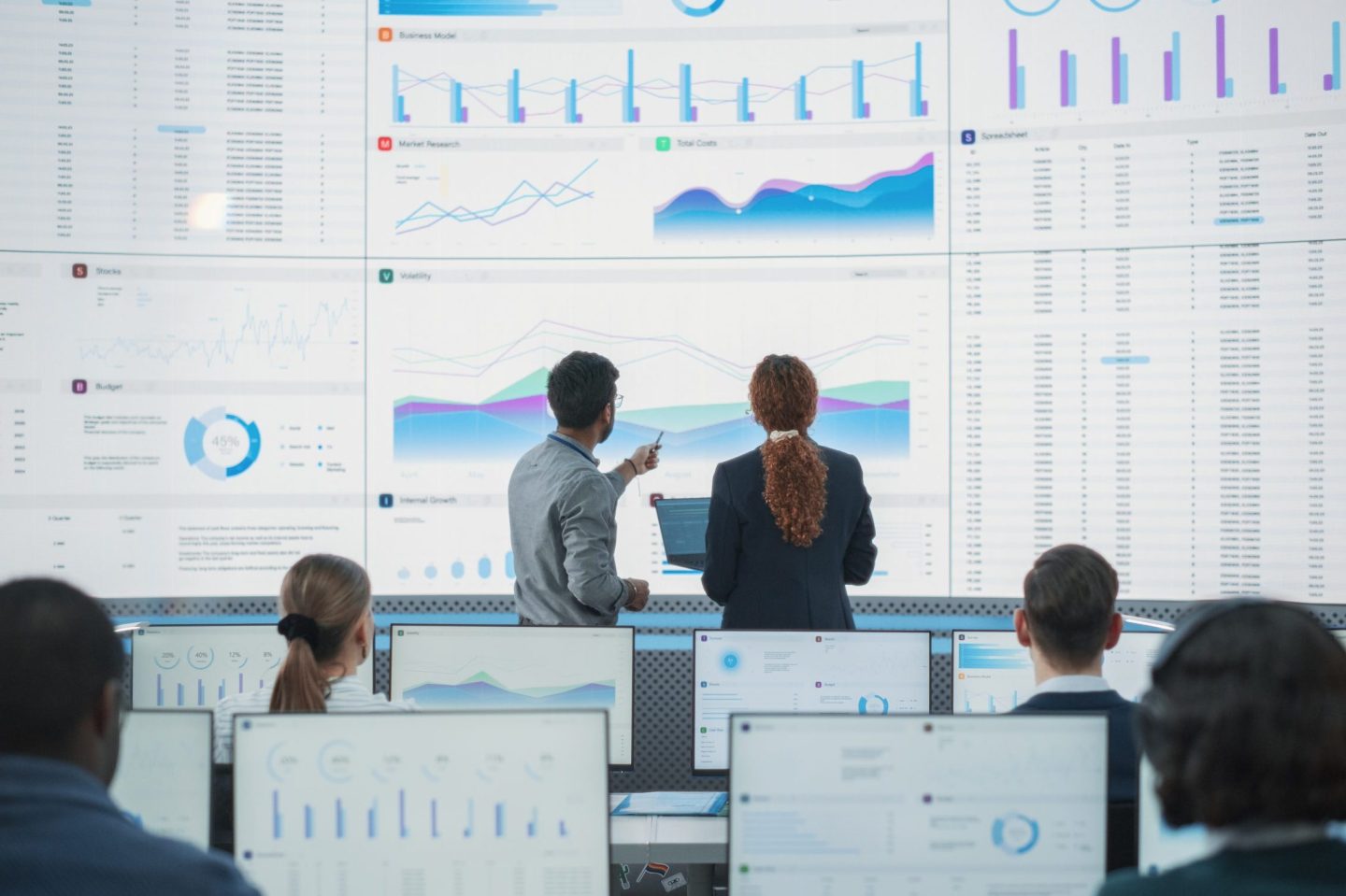 Wide Shot of Diverse Male And Female Data Analysts Discussing Graphs And Reports On Big Digital Screen In Monitoring Office. Multiethnic Employees Working On Desktop Computers for AI Company.