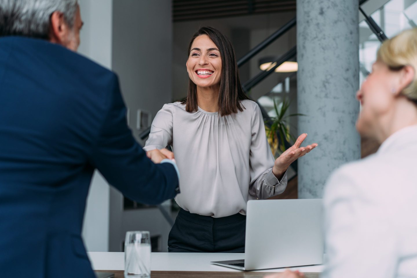 A young woman in business professional attire shakes hands with a man wearing a suit and sitting across a table from her.