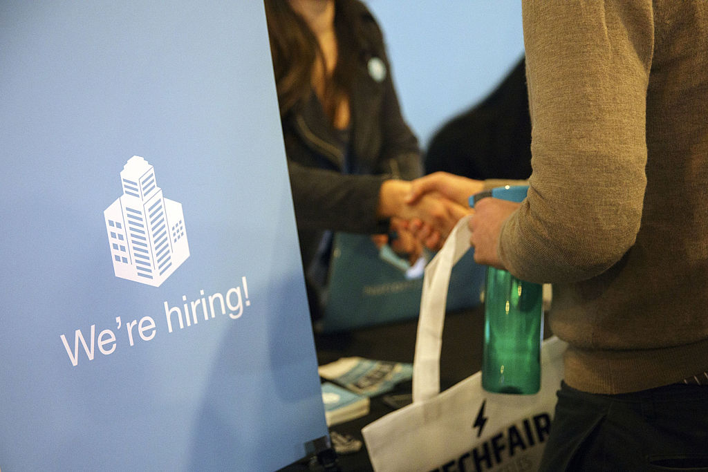 A sign saying "We’re Hiring" is displayed as a job seeker shakes hands with a representative during the TechFair L.A. job fair in Los Angeles, as seen in January 2017. (Photographer: Patrick T. Fallon—Bloomberg/Getty Images)