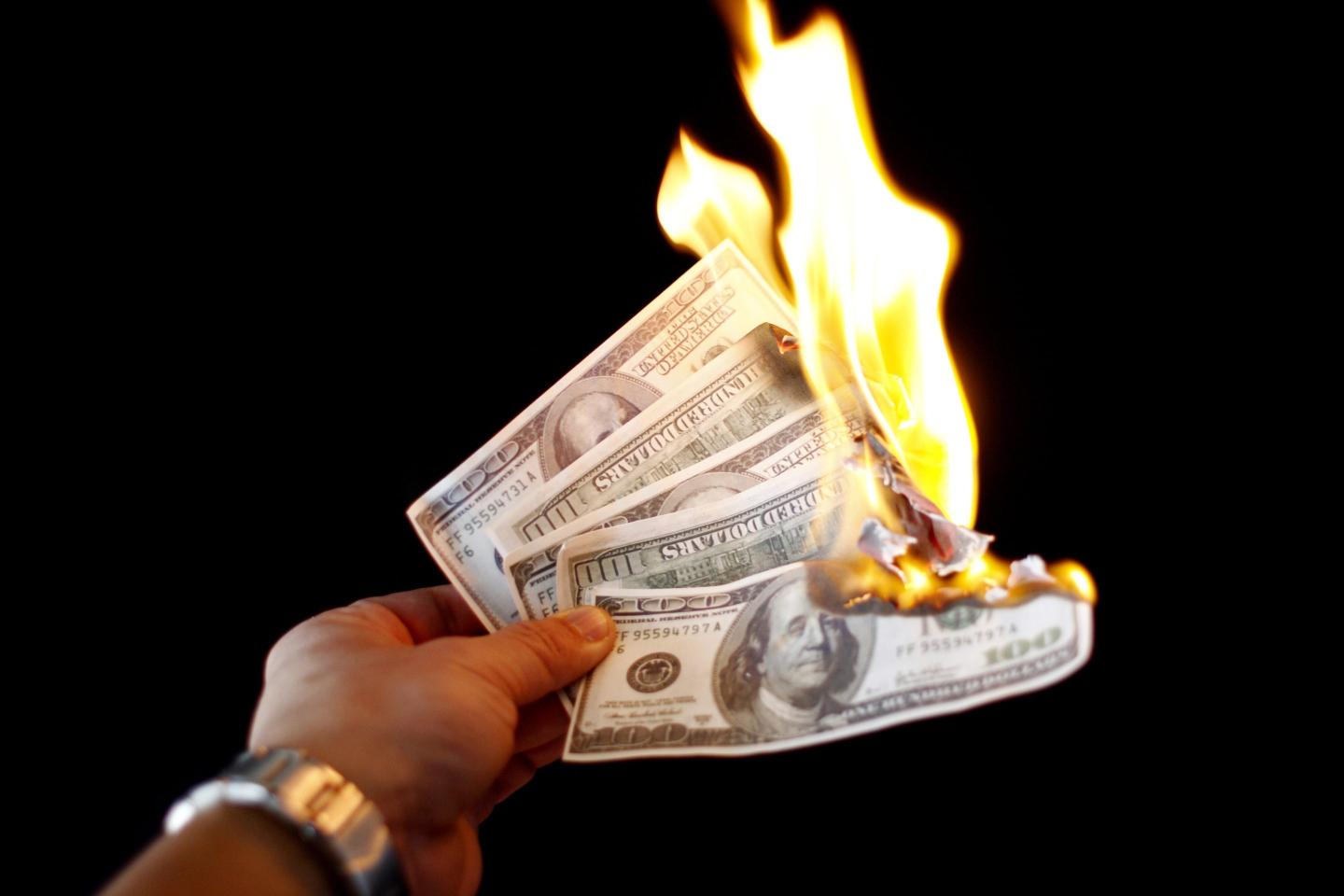 Stock photo of a hand holding several one hundred dollar bills, which are on fire.