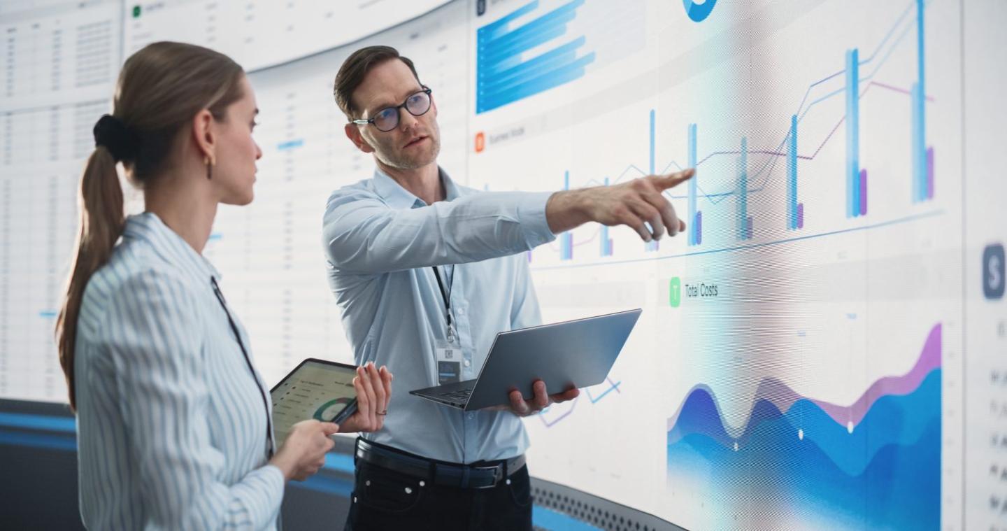 Man holding a laptop points to a graph while women with a tablet watches