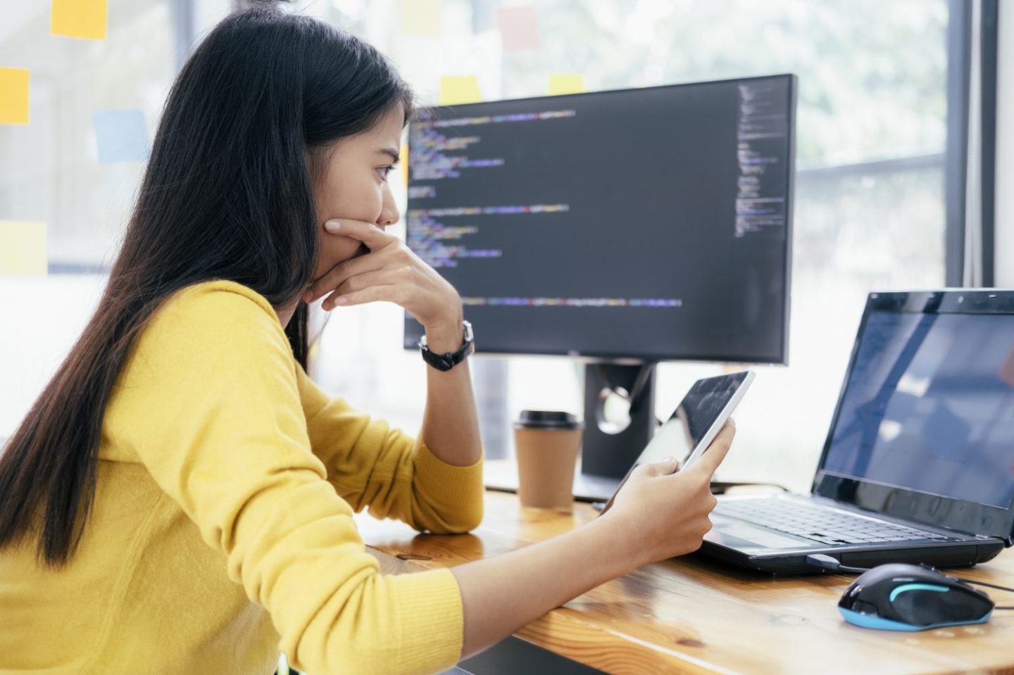 Woman holds a tablet and sits at a desk that has code open on a monitor and laptop