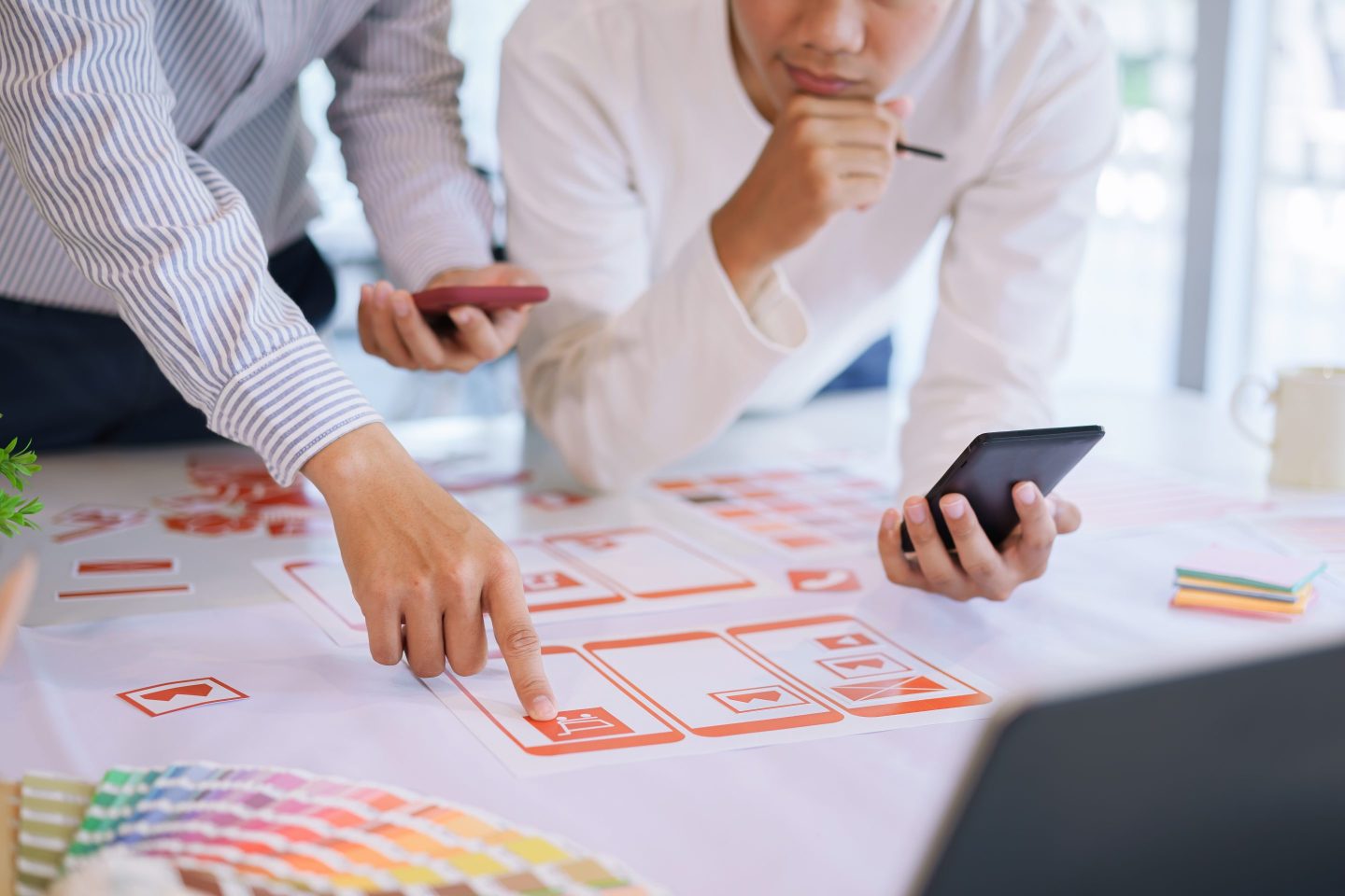 With phones in their left hands, UX designers work on a project on the table in front of them.