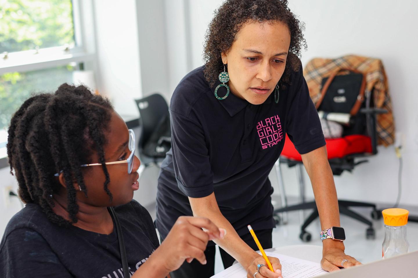 Girl with glasses is helped with an assignment by a woman with a Black Girls Code staff shirt on
