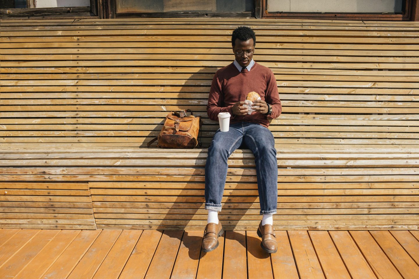 oung man sitting on wooden bench, eating hamburger, drinking coffee