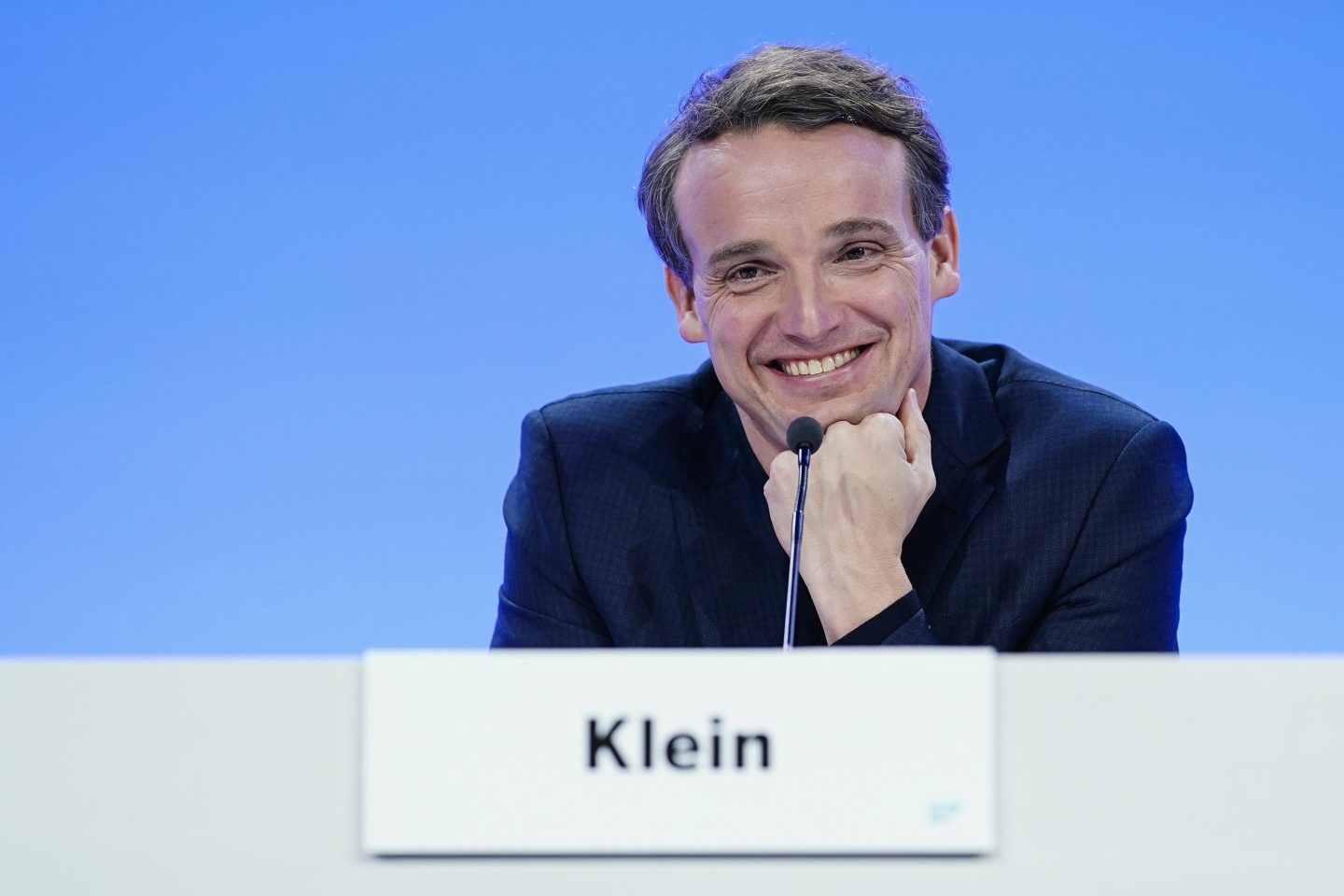 A blonde-haired man smiling and wearing a suit–sitting behind a mic.