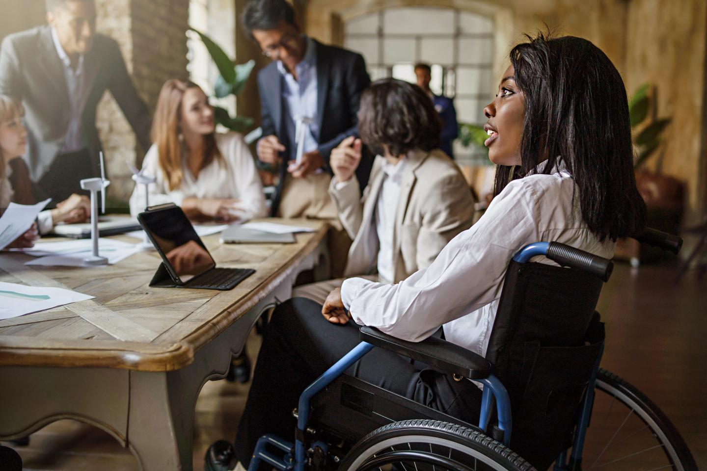 Picture of several businesspeople sitting at a board room-style table. At the center of the shot is a woman in a wheelchair.