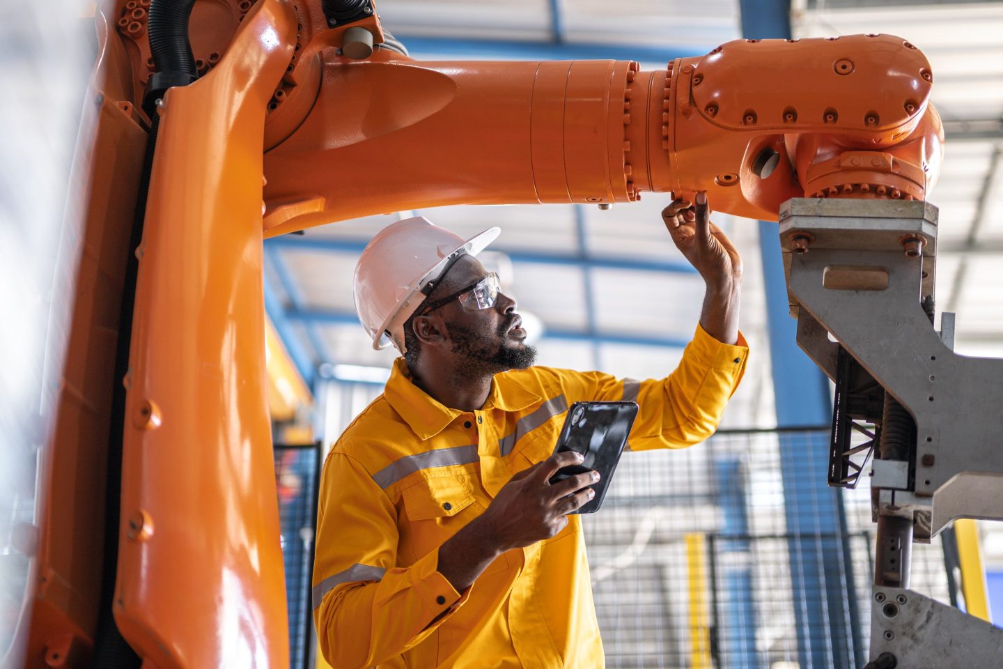 A man wearing a hard hat and personal protective equipment inspects a robotic arm in a factory.