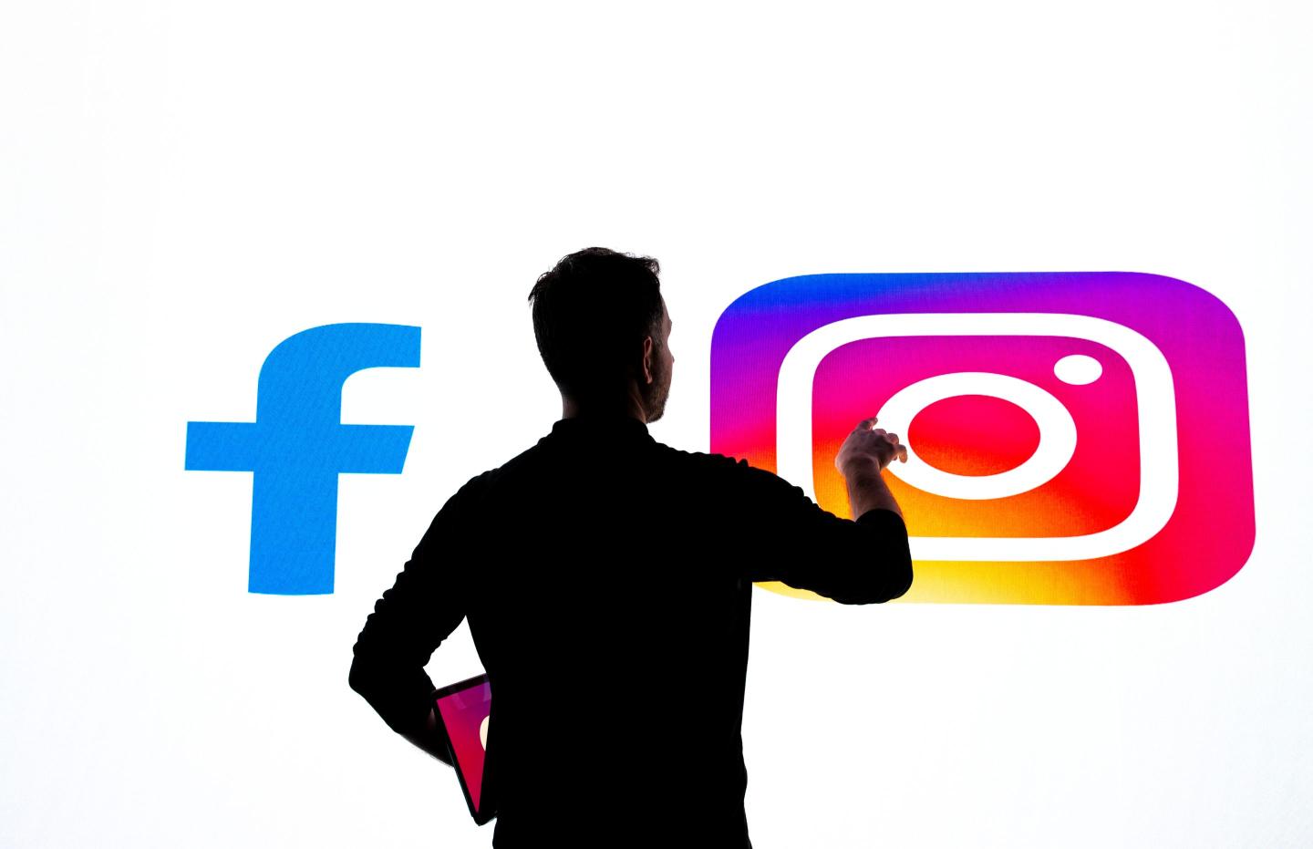 In this photo illustration, logos of 'Instagram' and 'Facebook' are displayed on a wide screen with a person silhoutte in the foreground
