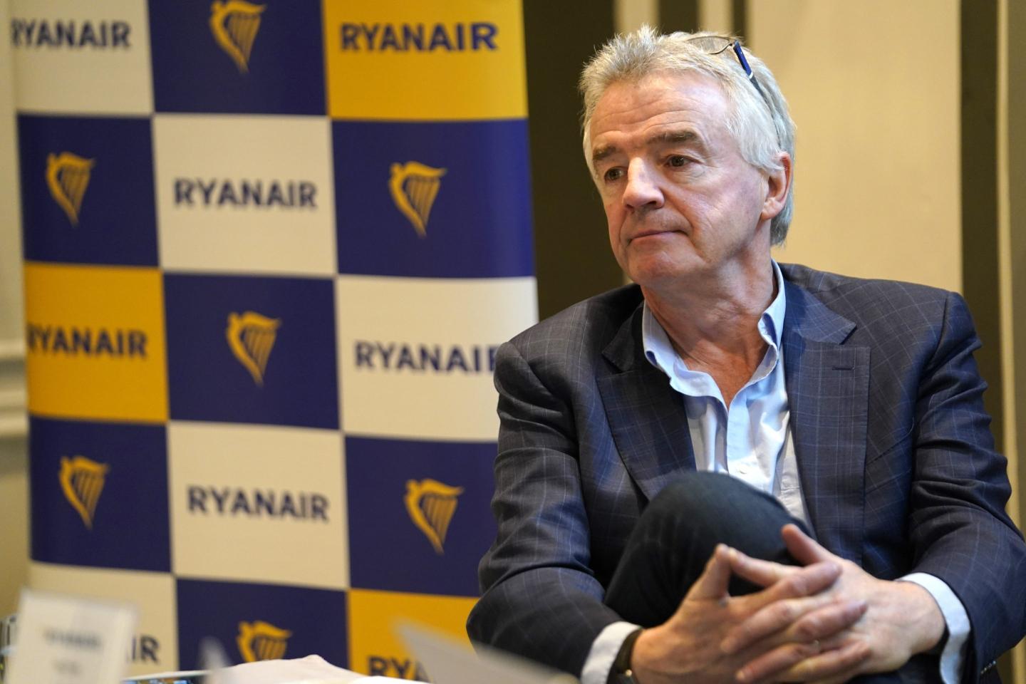 Ryanair CEO Michael O'Leary is sitting cross-legged in front of a checkered Ryanair background.