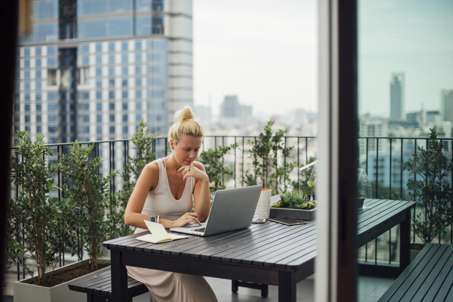 A woman uses a laptop while sitting on a balcony. There is a city landscape behind her.