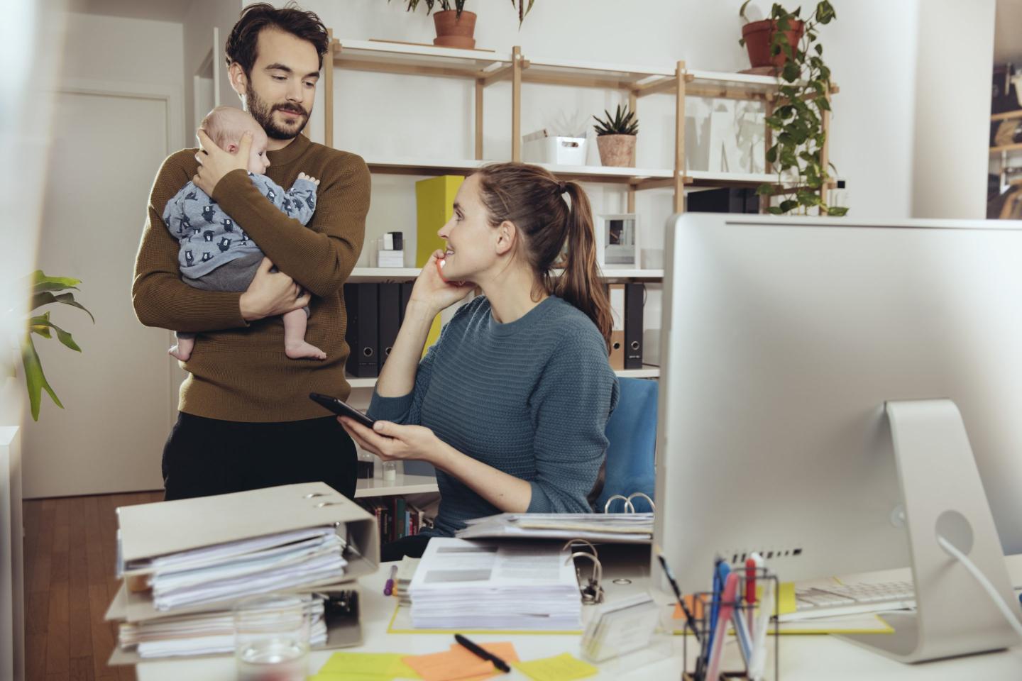 Smiling mother at desk looking at father holding baby in home office.