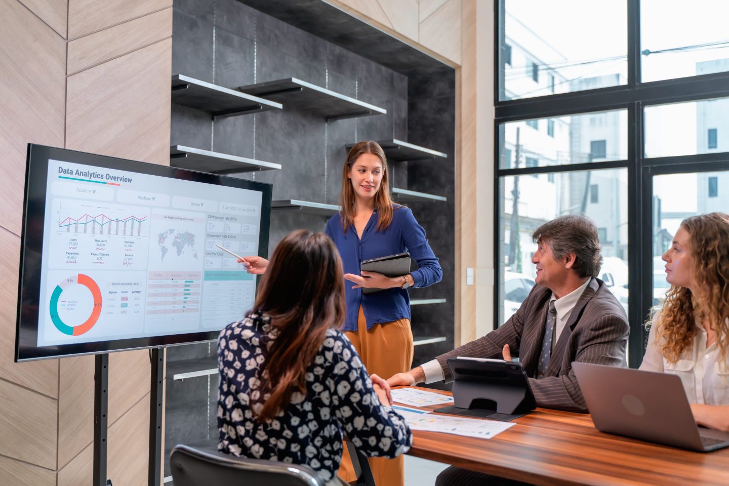 Woman presents data that is displayed on a TV to a group sitting at a conference table.