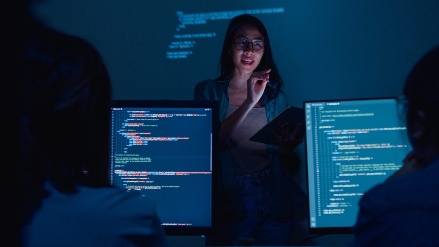 Young woman teacher lectures software developers in dark room.