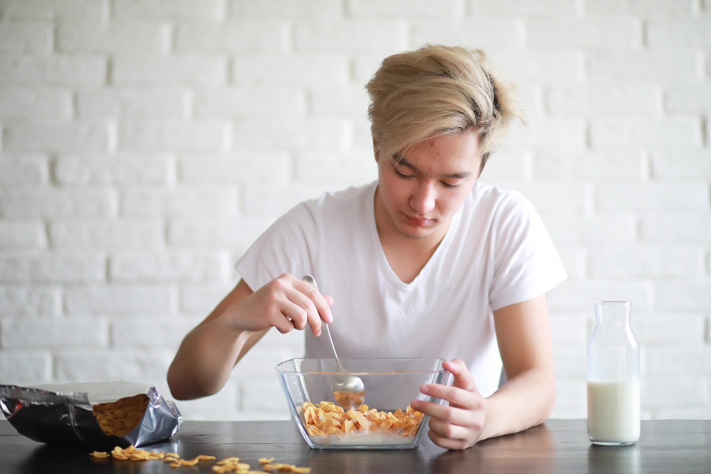 A young man in front of a white brick background eats corn flakes from a glass bowl.