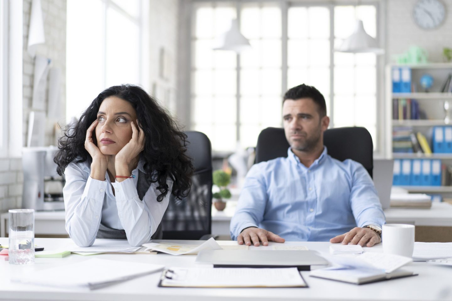 Two coworkers sit at a desk in an office. Both look upset.