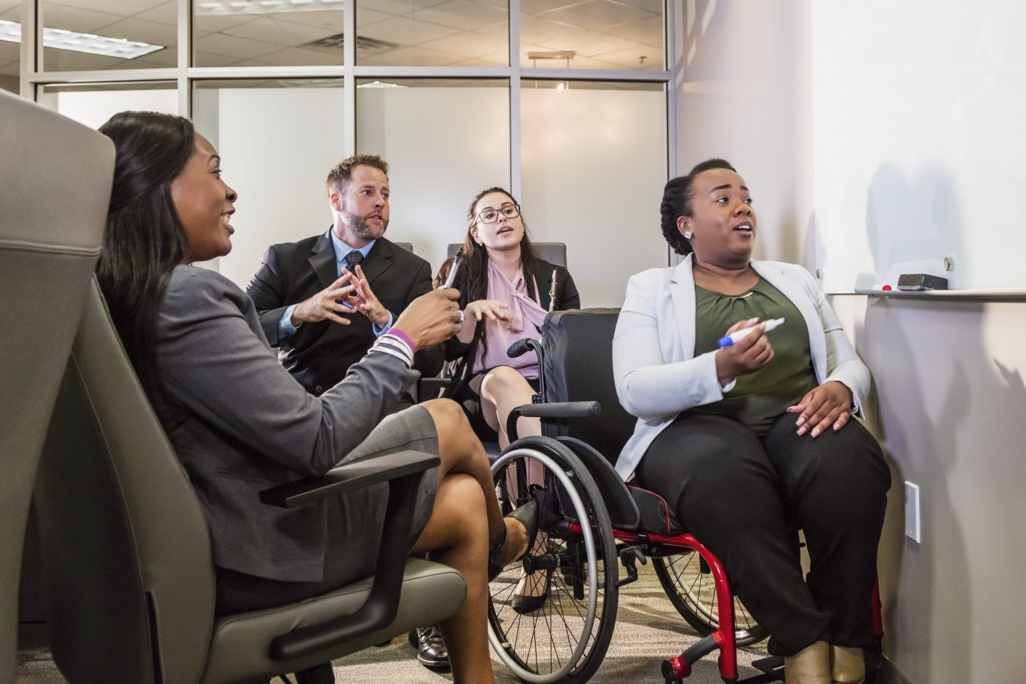 A group of four business people meeting in an office boardroom. A woman who uses a wheelchair is presenting at a white board.