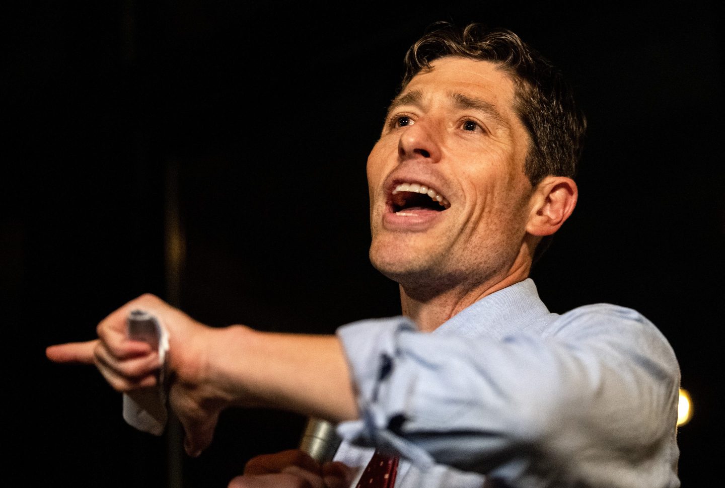Jacob Frey is wearing a blue dress shirt and gesturing with a microphone in his hand.