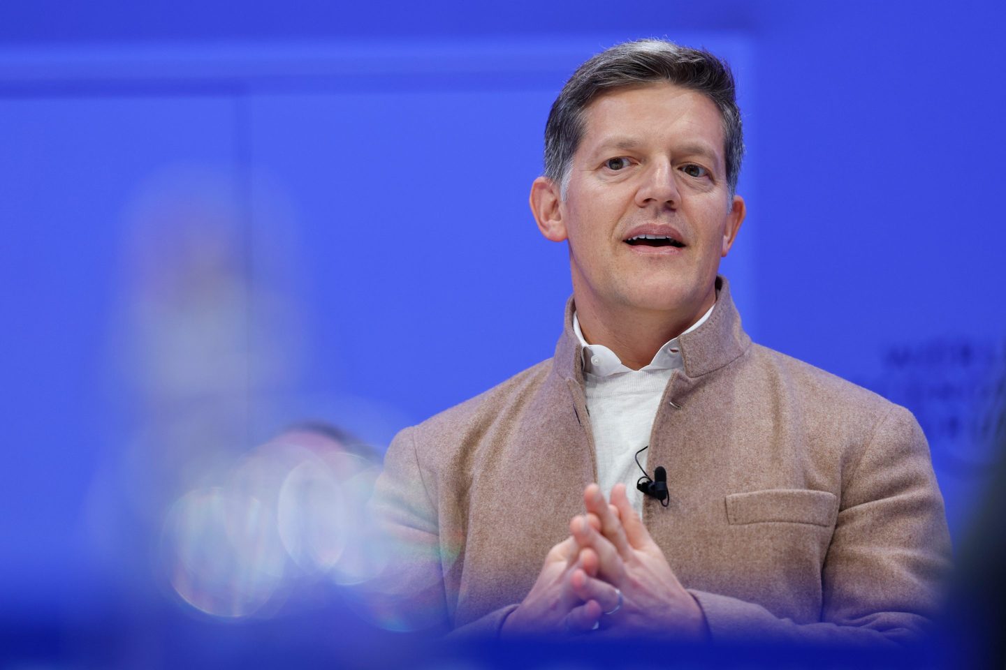Roelof Botha is wearing a tan suit and speaking in front of a blue-lit background.
