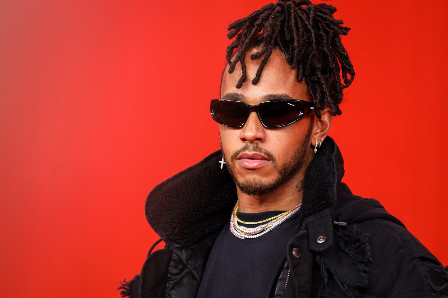Lewis Hamilton wears an all black outfit and dark sunglasses and stands in front of a red background.